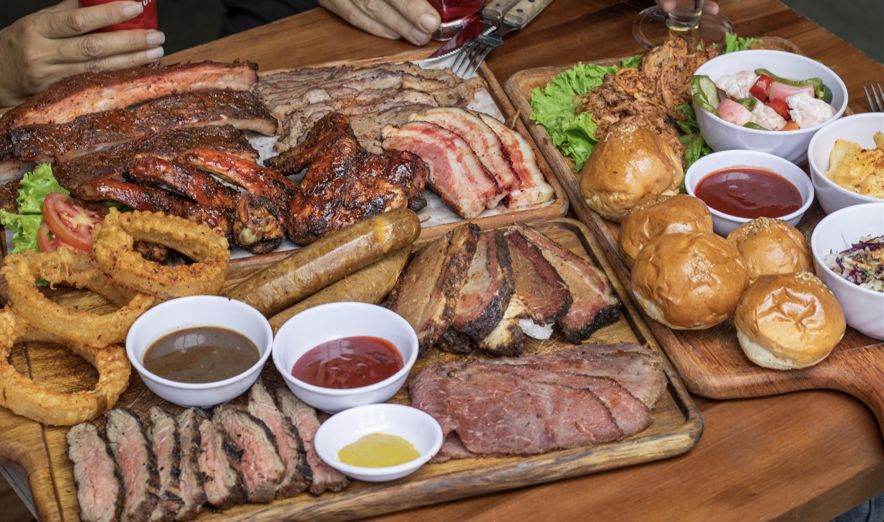 Sliced meat, ribs, chicken, sausages, onion rings, bread rolls and side dishes on wooden boards.