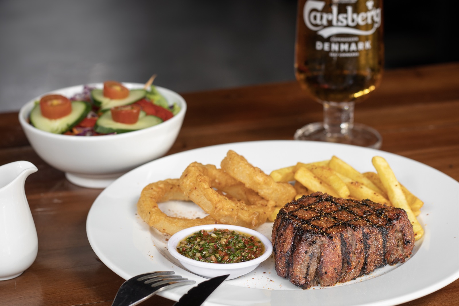 Steak with fries, onion rings, salad and sauce.